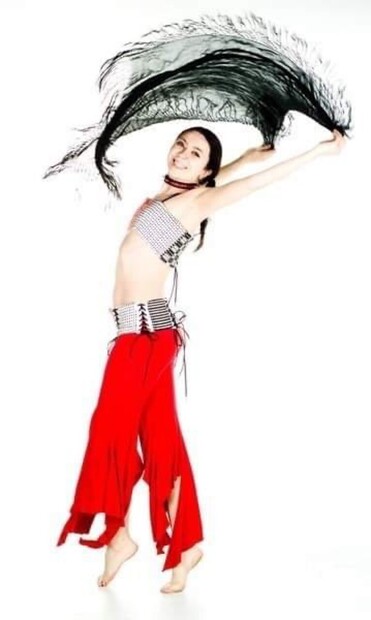 I’m wearing bright red flared pants, a black, white, and red handwoven corset belt, and a matching bandeau top. I’m swinging a black fringed scarf overhead and am wearing a black and red spiked collar. 