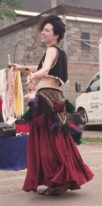 I’m smiling and dancing with my hair up as I play finger cymbals. I’m wearing a black vest trimmed with coins and metallic rickrack, a triangular brown hip belt trimmed with chain and wool tassels, and a flowing dark red skirt. 